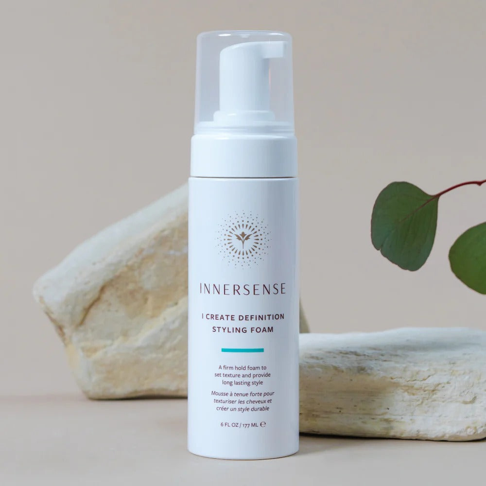 White Innersense I Create Definition Styling Foam bottle with a clear pump, set against natural rocks and a green leaf.