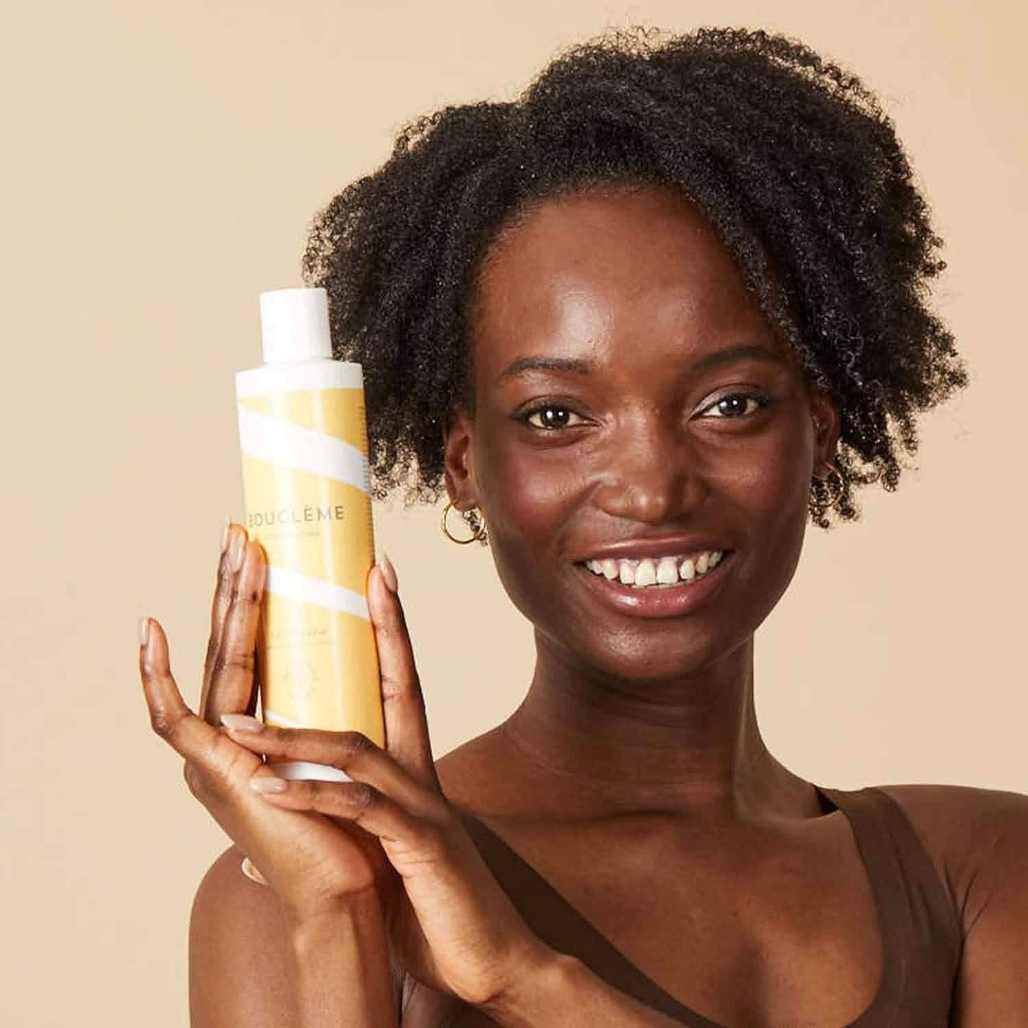 Smiling woman with curly hair holding a yellow Bouclème Curl Conditioner bottle with white stripes.
