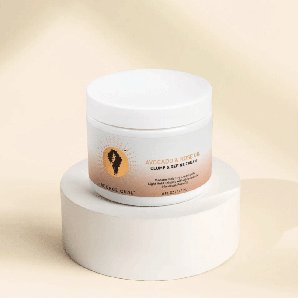 White jar of Bounce Curl Avocado & Rose Oil Clump & Define Cream with a sunburst logo, on a white pedestal.