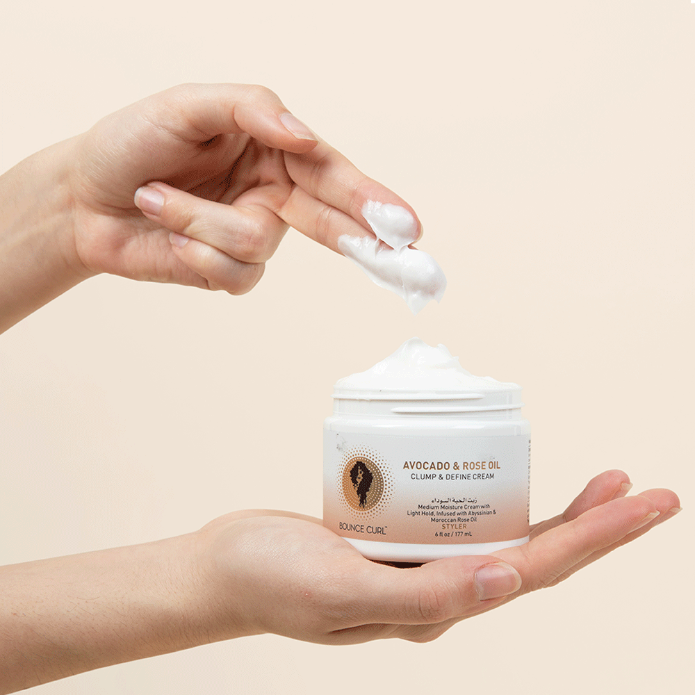 Hands holding a white jar of Bounce Curl Avocado & Rose Oil Clump & Define Cream with white product on fingers.