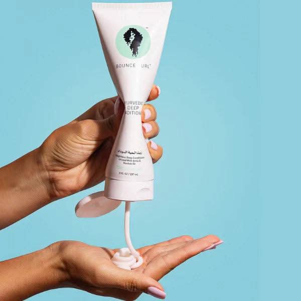 Hand squeezing Bounce Curl Ayurvedic Deep Conditioner from a white tube, dispensing pink cream into a palm.