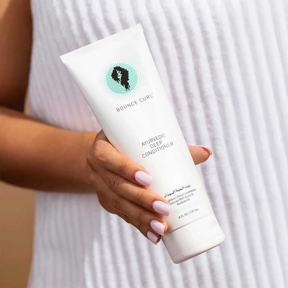 Hand holding a white Bounce Curl Ayurvedic Deep Conditioner tube with a teal and black logo.