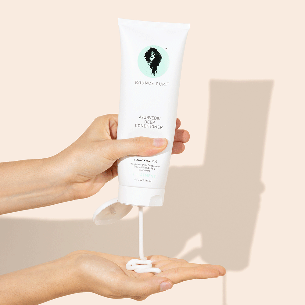 Hands squeezing white Bounce Curl Ayurvedic Deep Conditioner tube, dispensing creamy product into a palm.