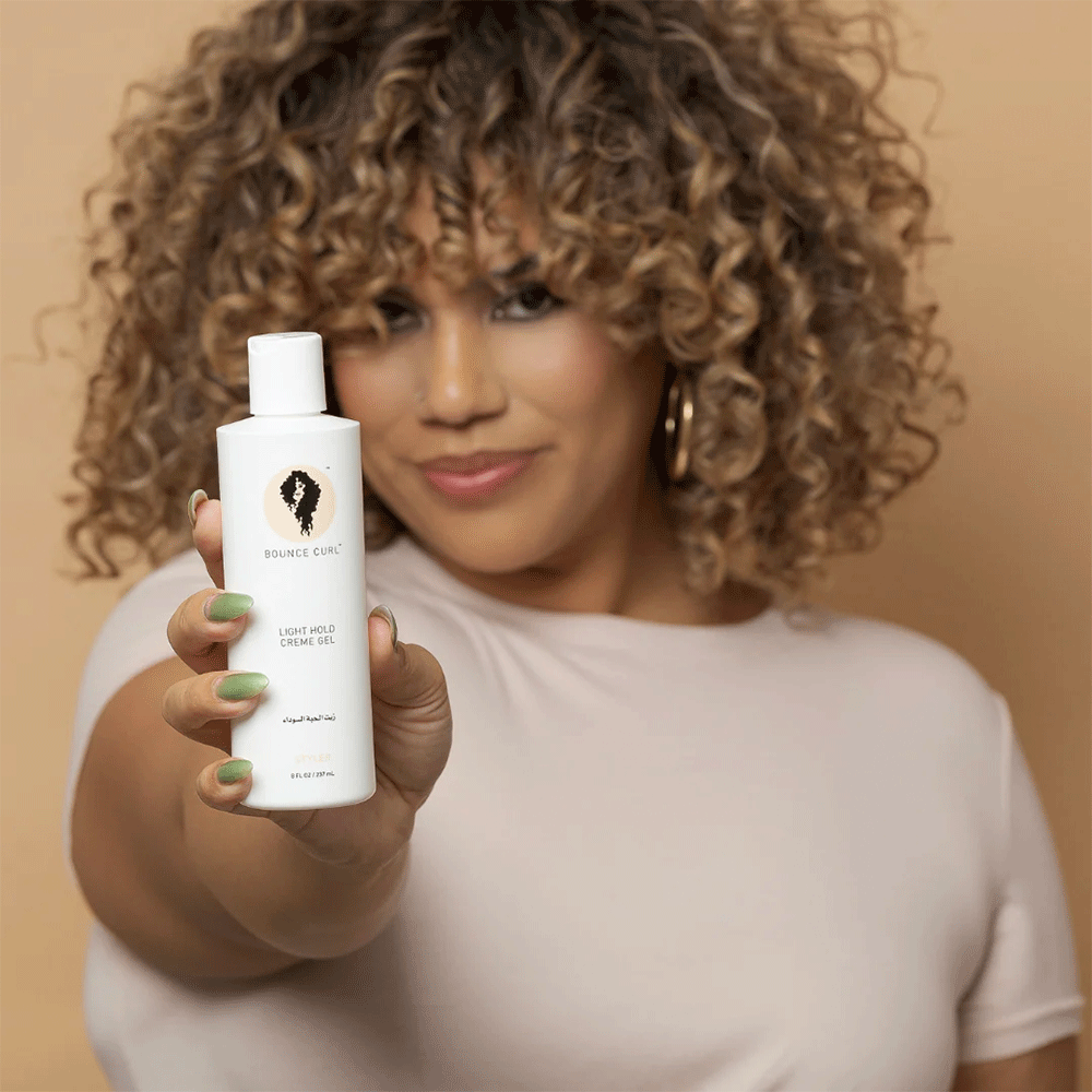 Woman with curly hair holding a white Bounce Curl Light Hold Cream Gel bottle.