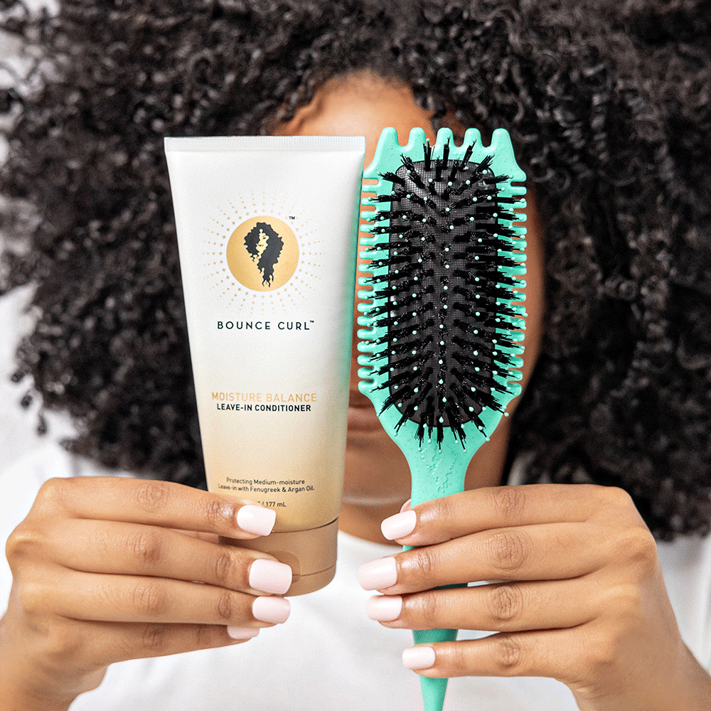Bounce Curl Moisture Balance Leave-In Conditioner tube and a teal detangling brush held by a person with curly hair.
