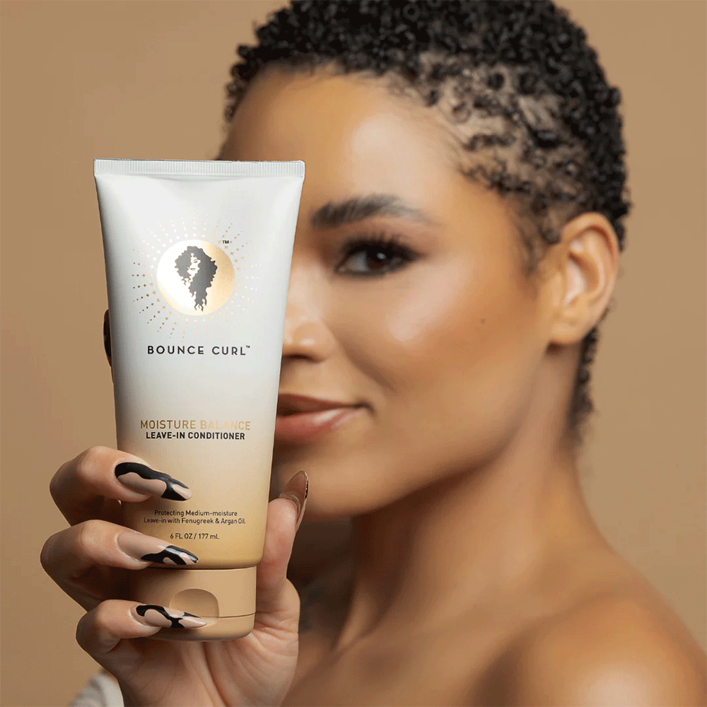 Woman with short curly hair holding a white and beige Bounce Curl Leave-In Conditioner tube.