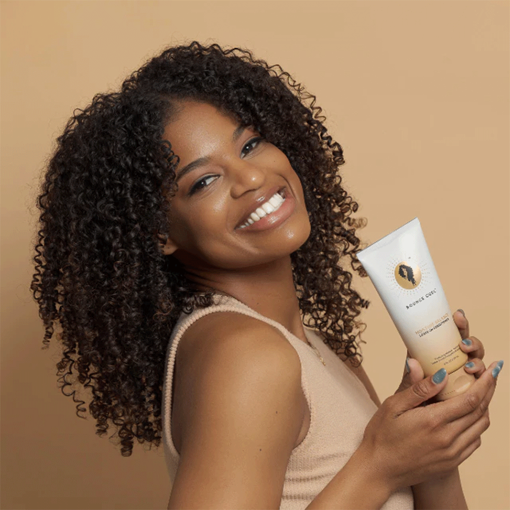 Smiling woman with curly hair holding a white Bounce Curl Moisture Balance Leave-In Conditioner tube.