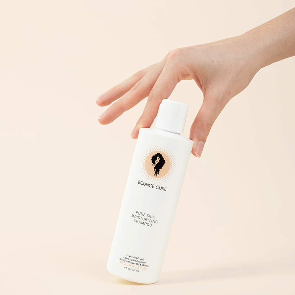 Hand holding a white Bounce Curl Pure Silk Moisturizing Shampoo bottle, 237ml, with a black and orange logo.
