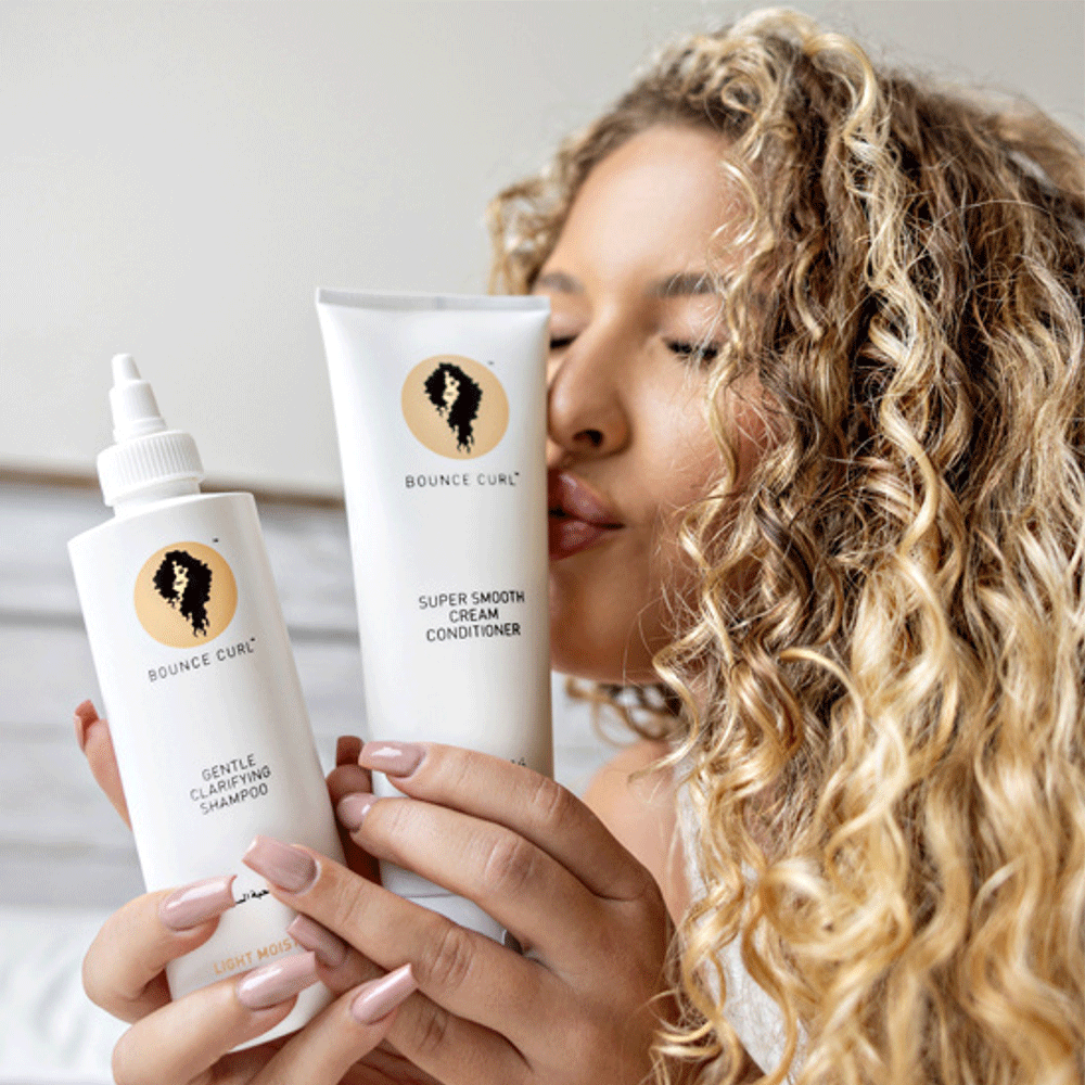 Woman with curly hair holding white Bounce Curl Super Smooth Cream Conditioner and Gentle Clarifying Shampoo.
