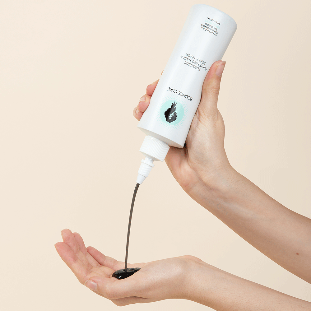 Hands squeezing a white Bounce Curl Turmeric Purifying Hair & Scalp Mask bottle, dispensing dark jelly into a palm.