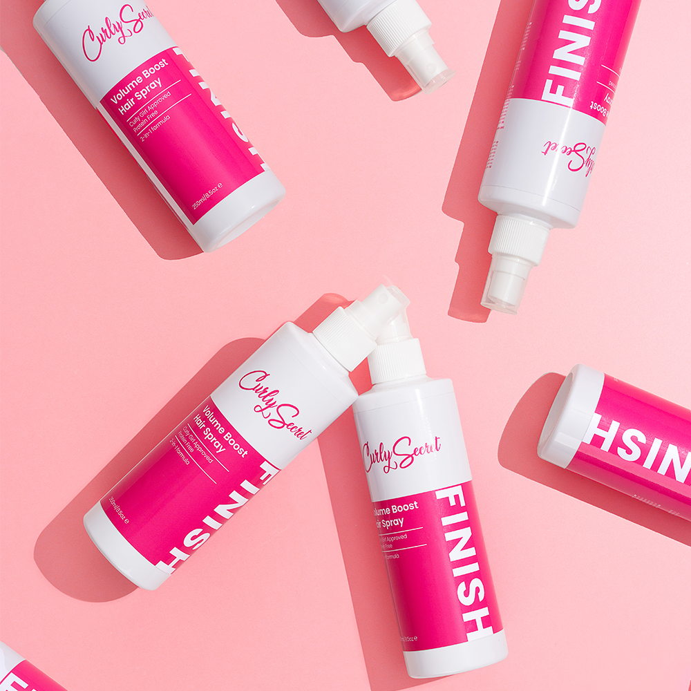 Multiple white and pink Curly Secret Volume Boost Hair Spray bottles with "FINISH" text on a pink background.