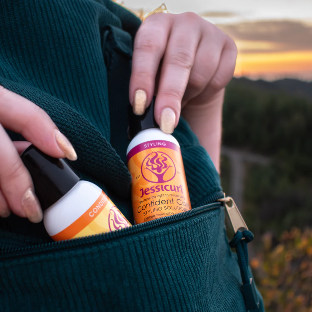 Hands placing two Jessicurl Confident Coils bottles, one orange and one yellow, into a green corduroy pocket.