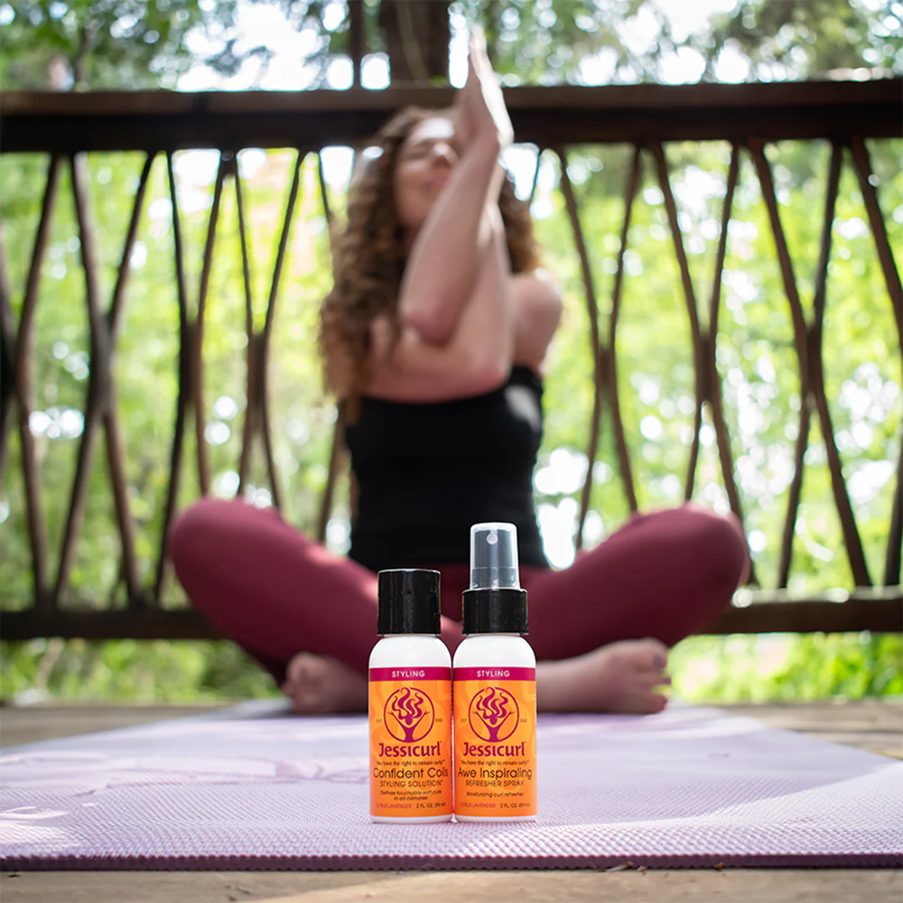 Two small Jessicurl styling product bottles, orange with black caps, on a yoga mat with a woman meditating in the background.