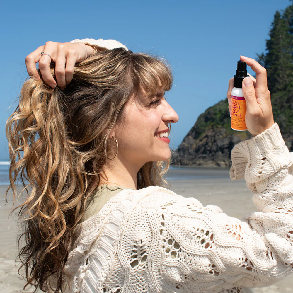 Woman with wavy hair on a beach holding an orange Jessicurl Gelebration Spray bottle.