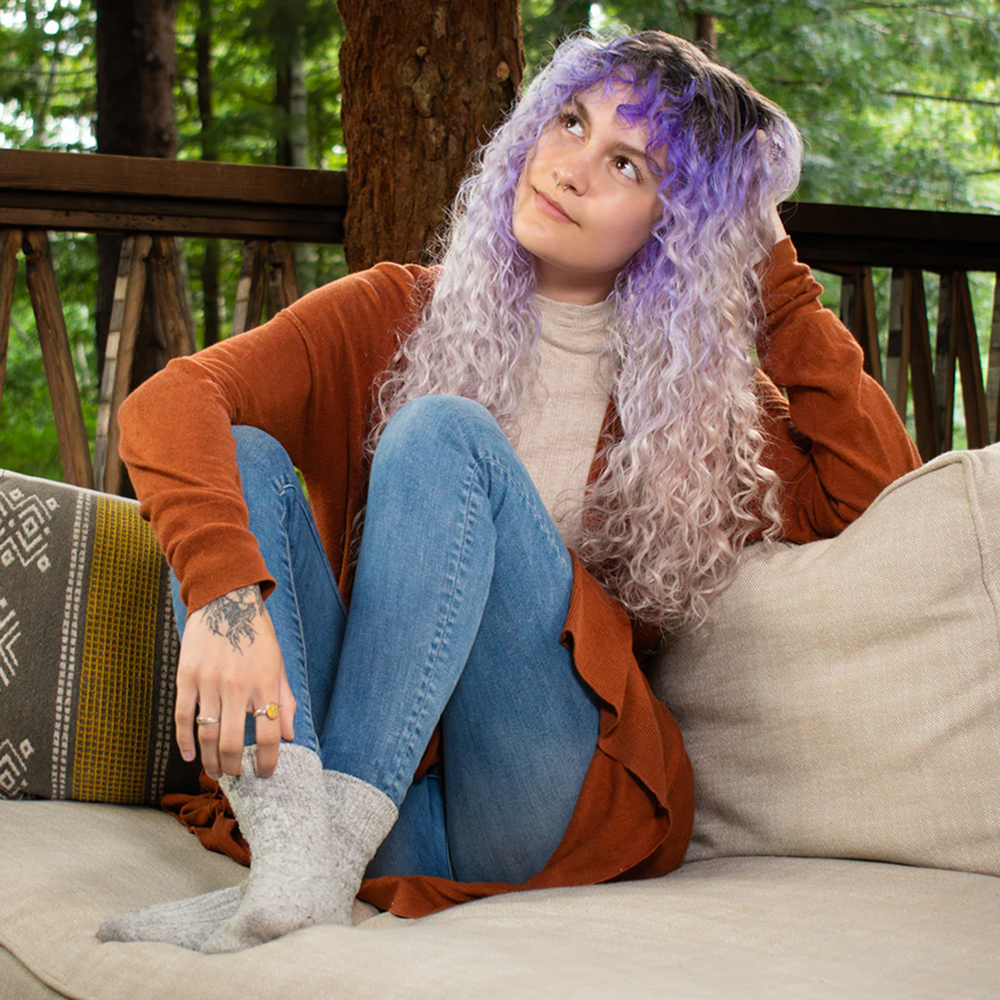 Young woman with long, wavy purple and blonde hair, wearing a rust cardigan and jeans, sitting on a patio couch.