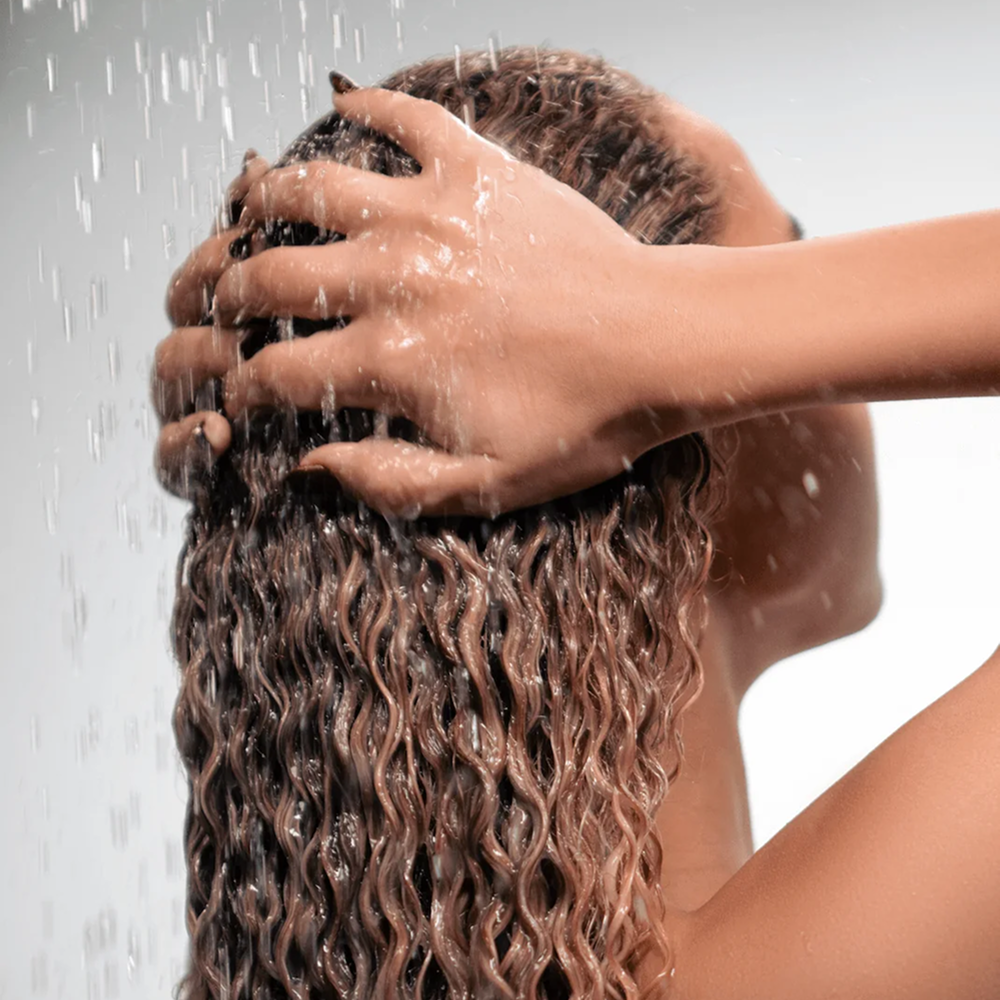 Person with light brown curly hair washing it under a shower, hands massaging scalp.